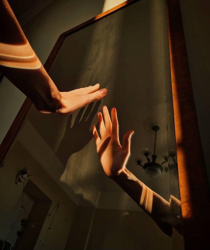 Dos manos tocan un espejo en una habitación iluminada por la luz solar que entra por la ventana.